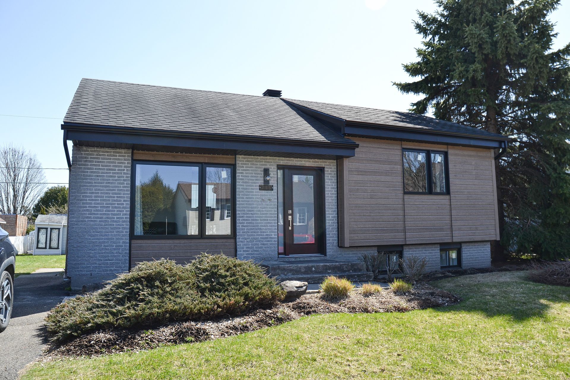 Maison de plain-pied avec briques grises et revêtement en bois, toit noir et cour avant avec herbe verte.