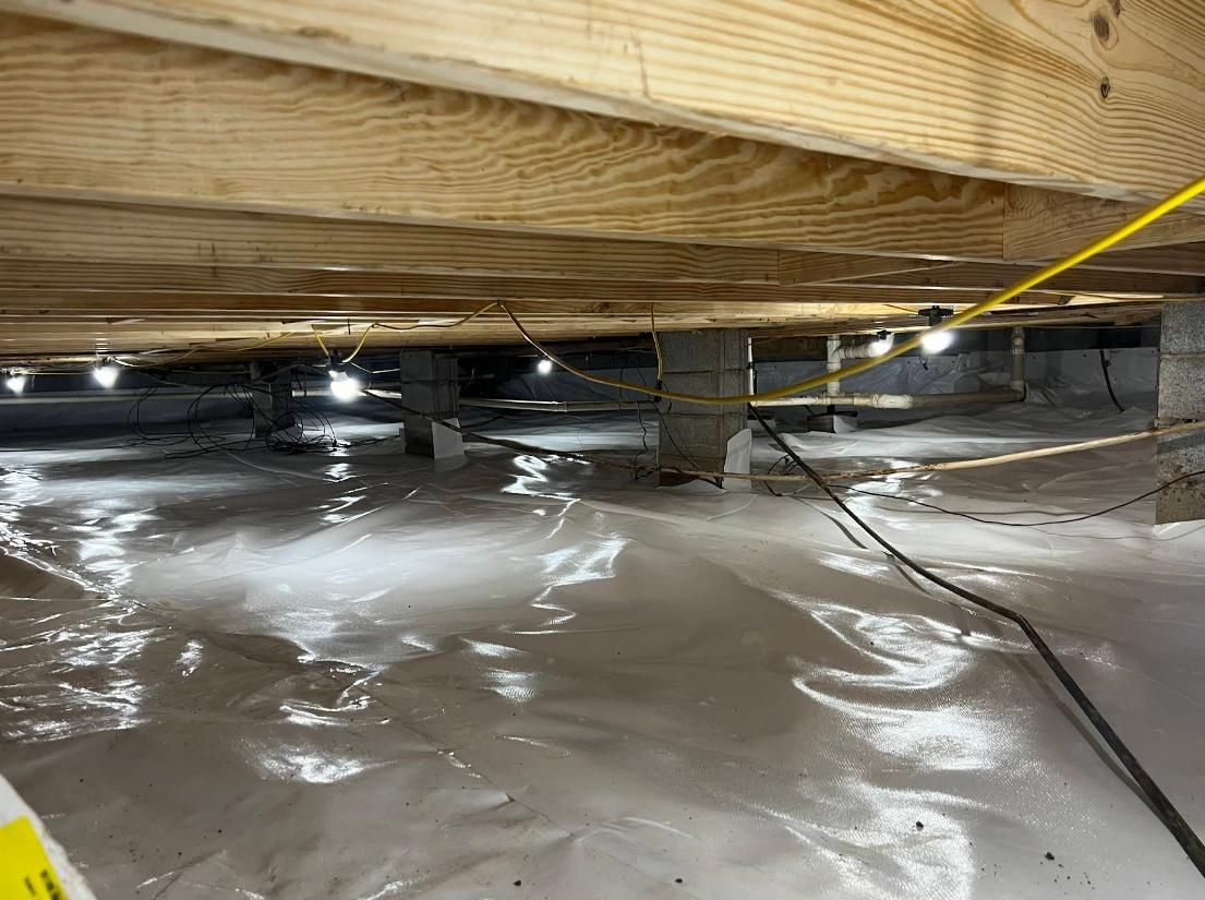 View of a crawlspace with white vapor barrier on the ground, wood beams above, and support columns. Lighting illuminates the area.