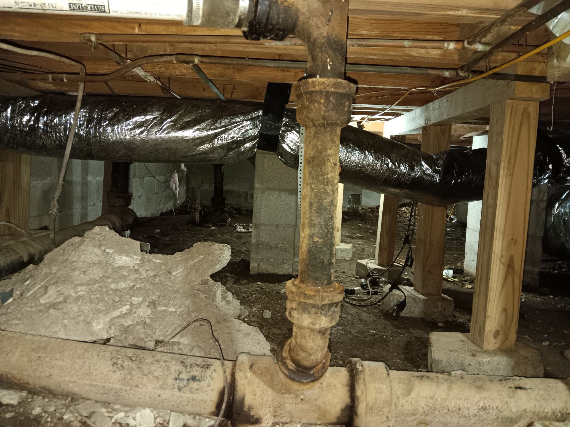 Underneath a house, aged pipes and ductwork among cinder blocks and wood supports.