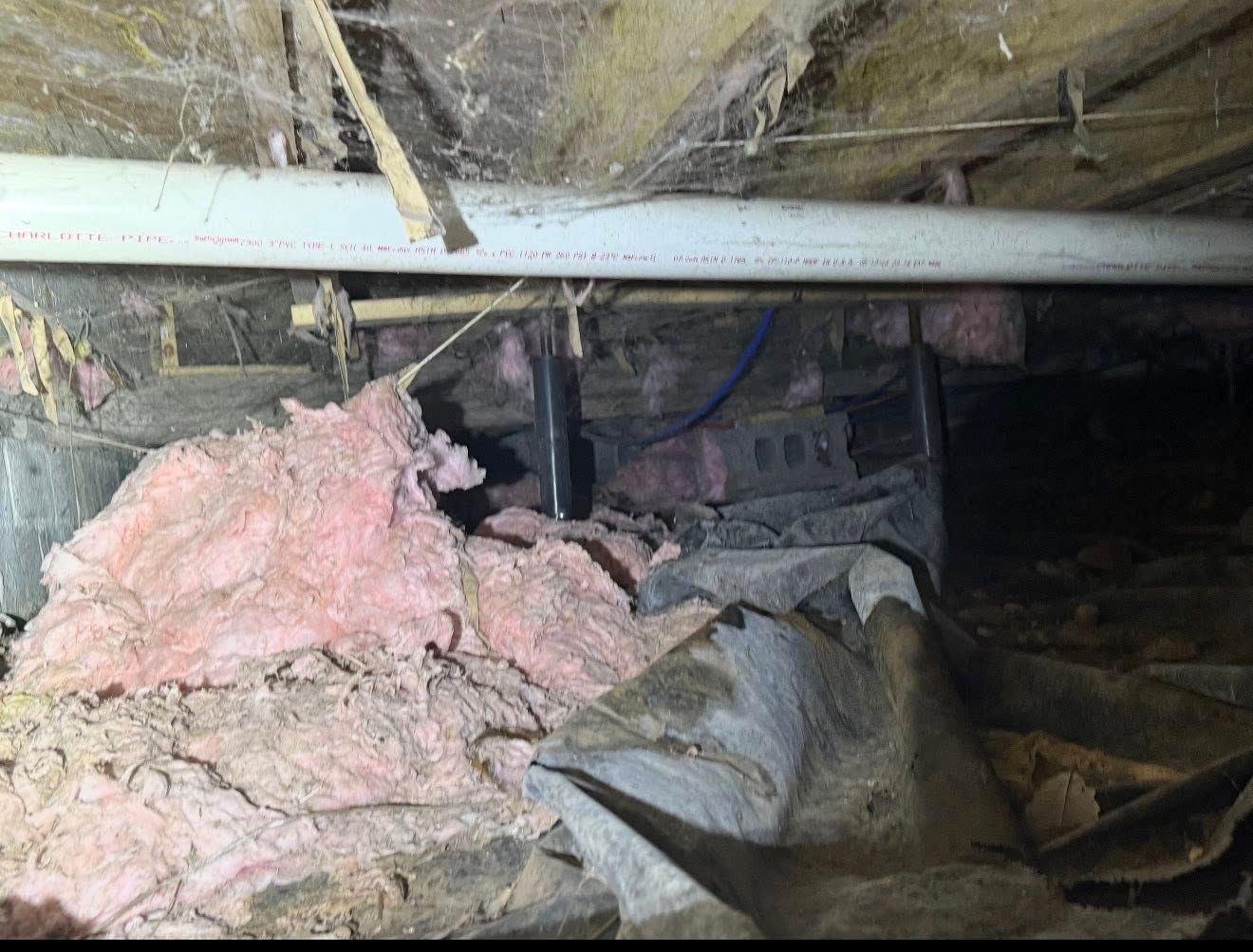 Crawl space with pink insulation, pipes, and debris. Dark, dusty setting with wood and a plastic sheet.