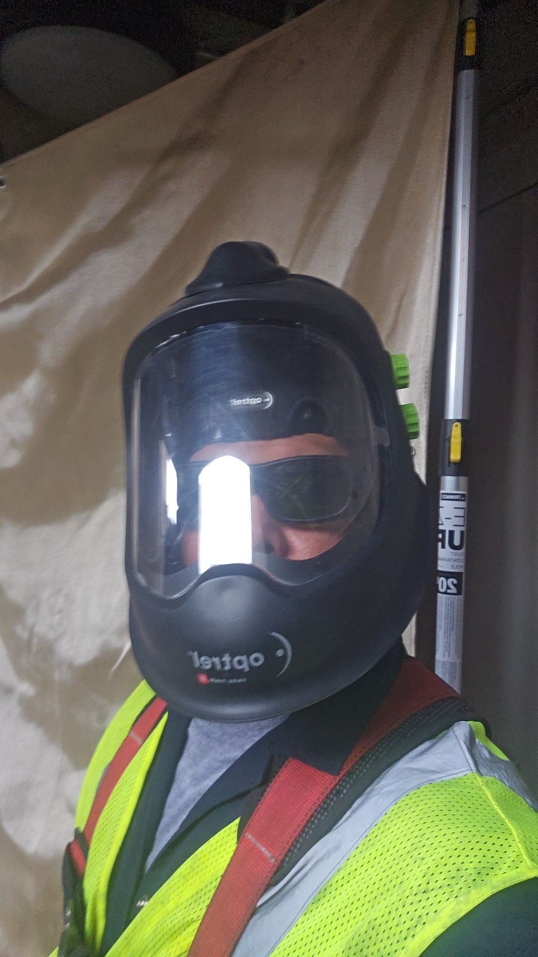 Person wearing a black respirator, safety vest, and carrying a pole indoors.