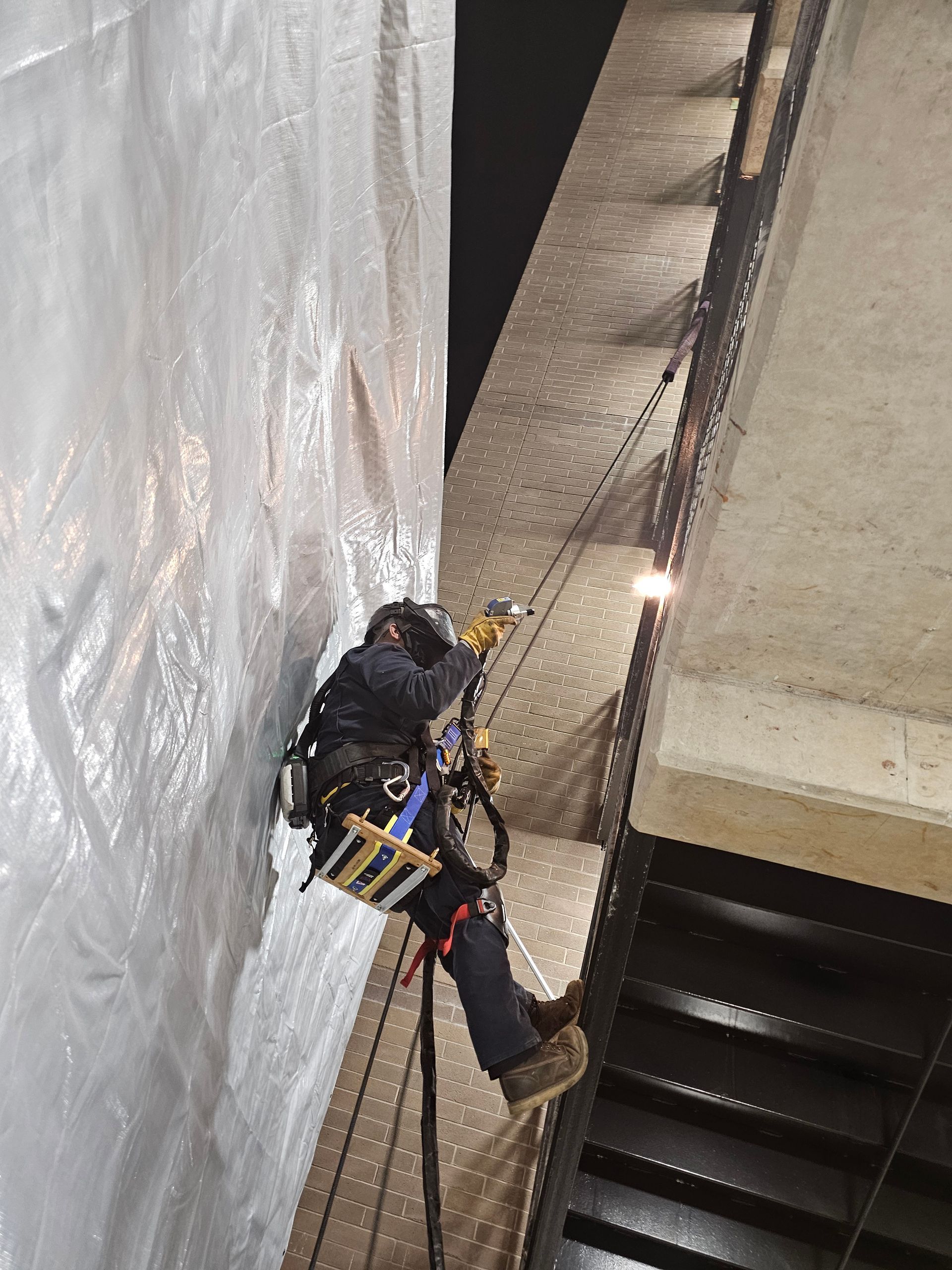 Rope access technician on a building inspecting a white tarp, wearing safety gear and tools.