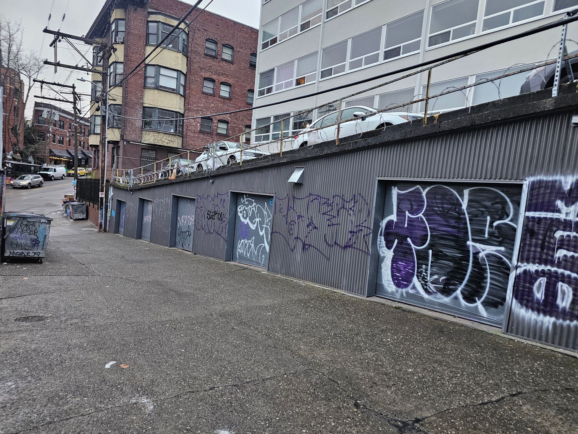 Graffiti-covered wall with doors along an alley. Buildings and parked cars in the background.