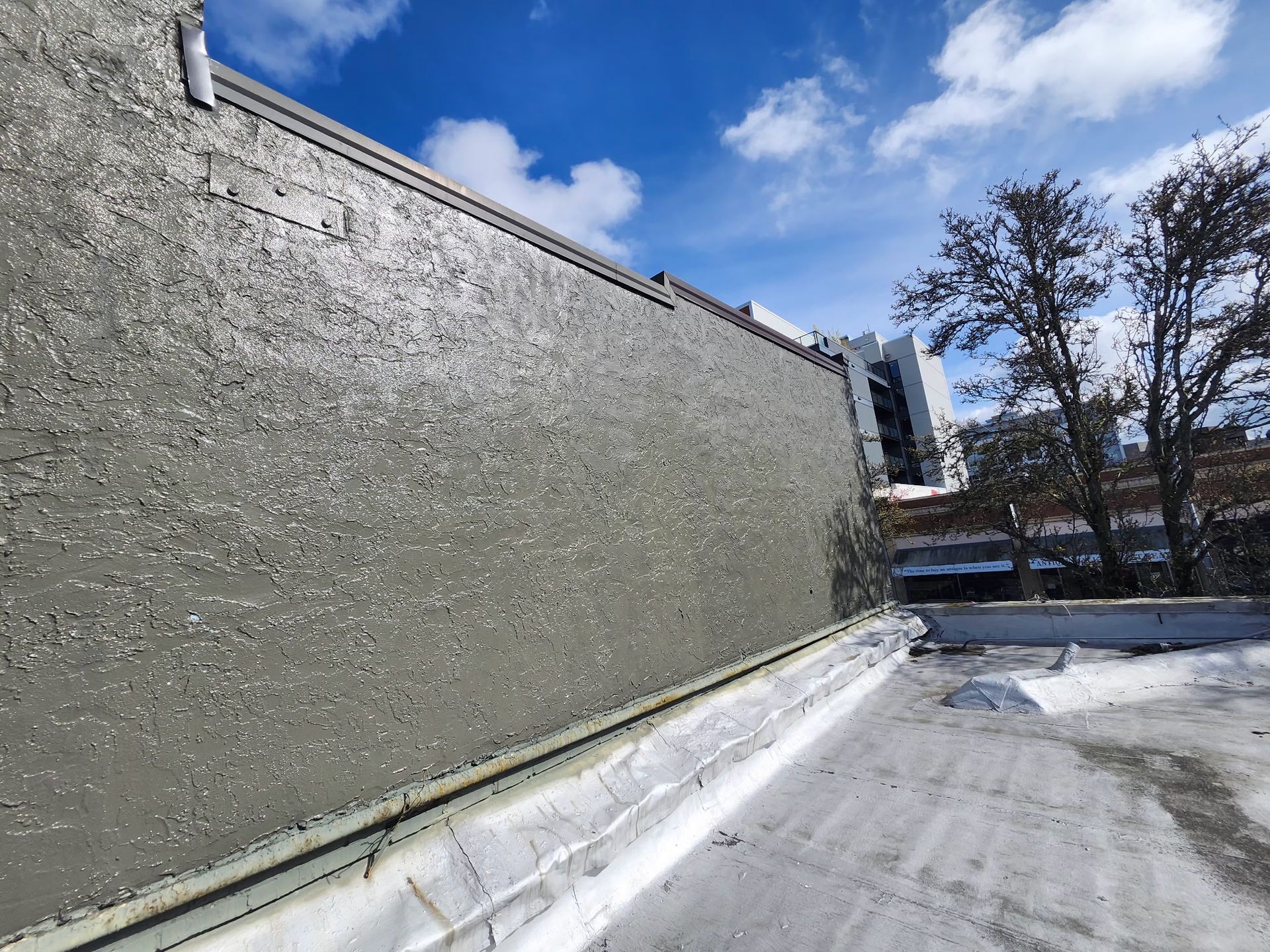 Gray wall with metallic trim on a rooftop under a blue sky with some trees.