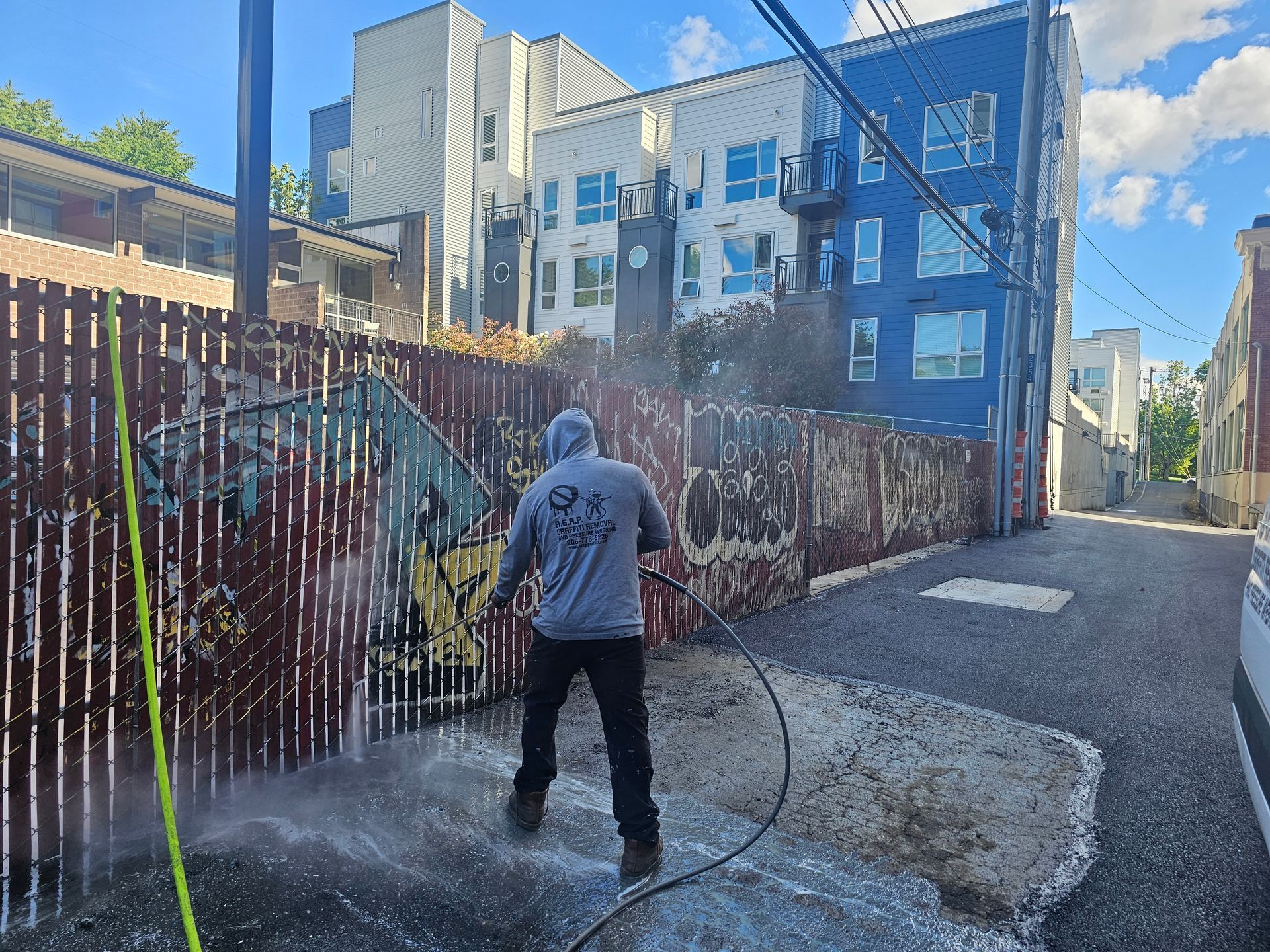 Person pressure washing a wooden fence with graffiti in an alleyway, near apartment buildings.