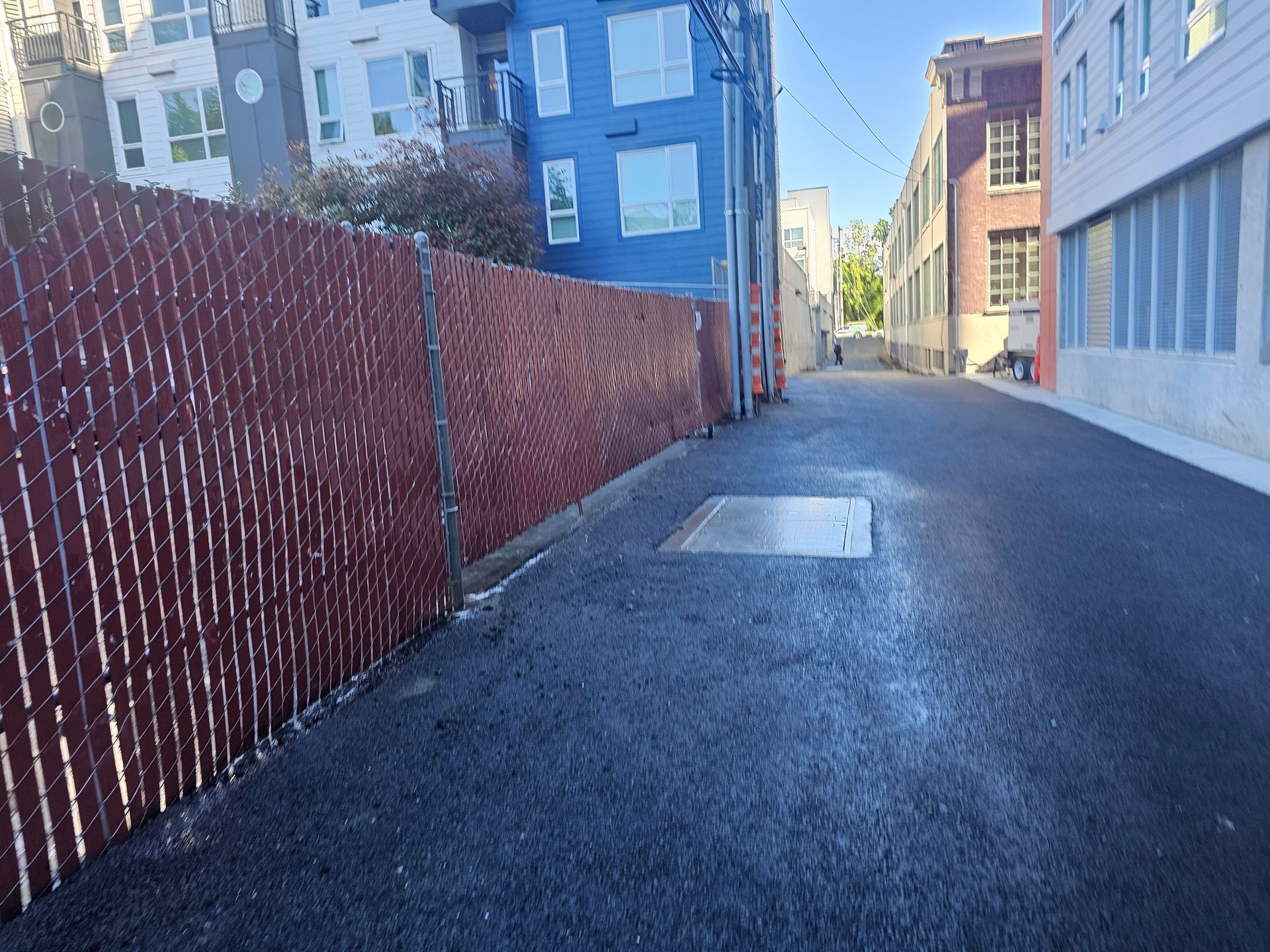 Alley with asphalt pavement, tall red fence on left, buildings on both sides, bright daylight.