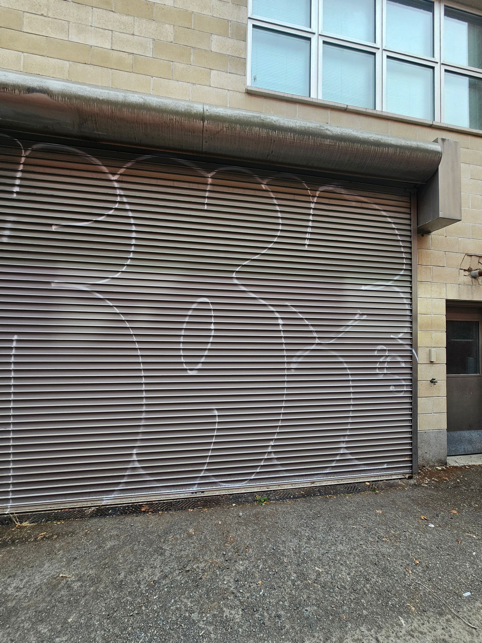 Gray metal roll-up door with white graffiti on a building with a window above.