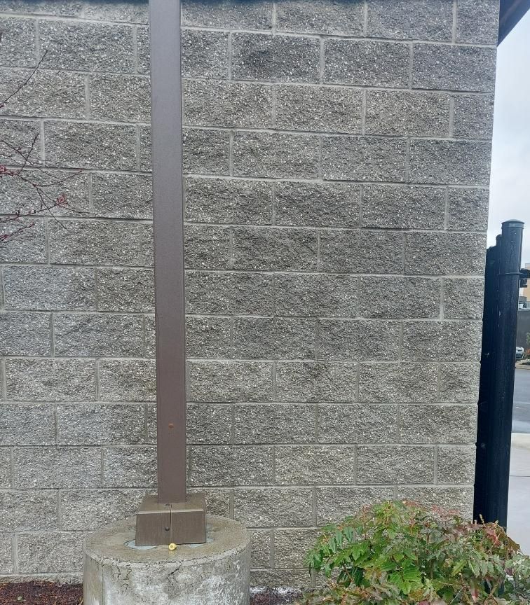 Gray cinder block wall with brown metal pole, on concrete base, with shrubs.