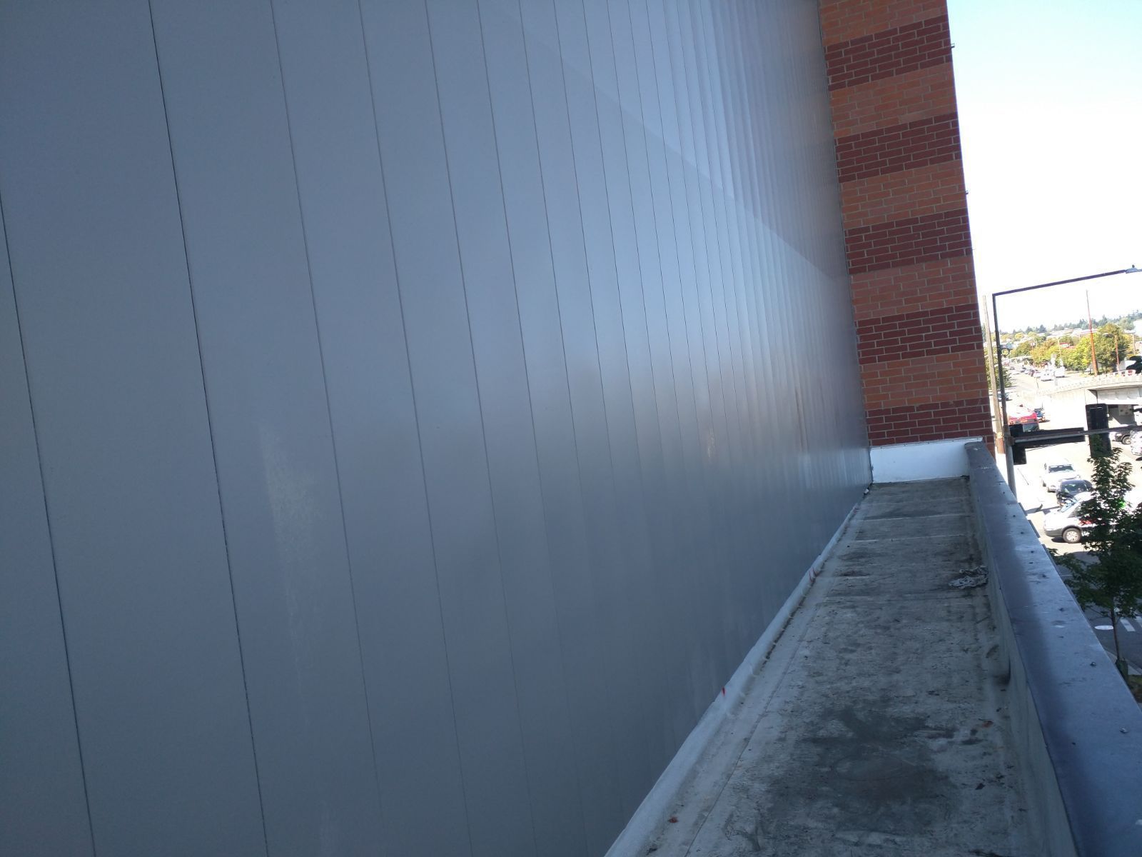Exterior view of a light gray wall, a red brick building, and a rooftop with concrete and a railing.