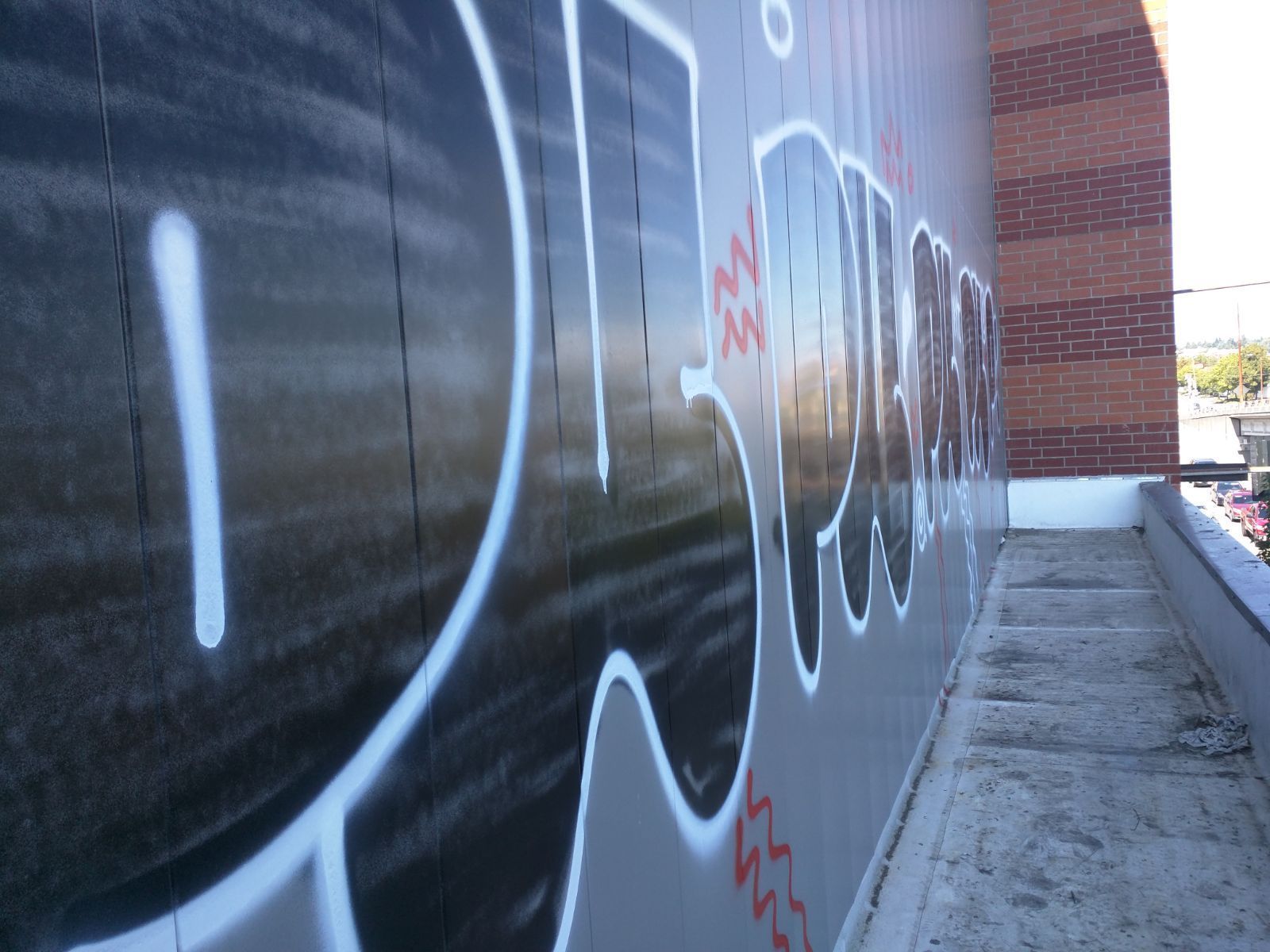 Graffiti on a light grey wall; white outline on black letters. Brick building in the background.