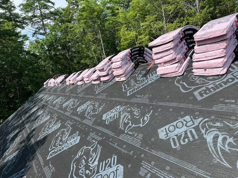 Stacks of pink roofing shingles on a roof covered with black underlayment, surrounded by trees.