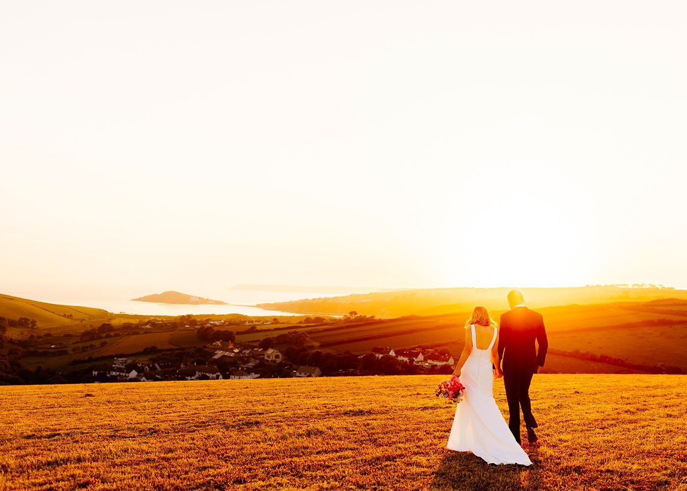 A bride and groom are walking in a field at sunset.