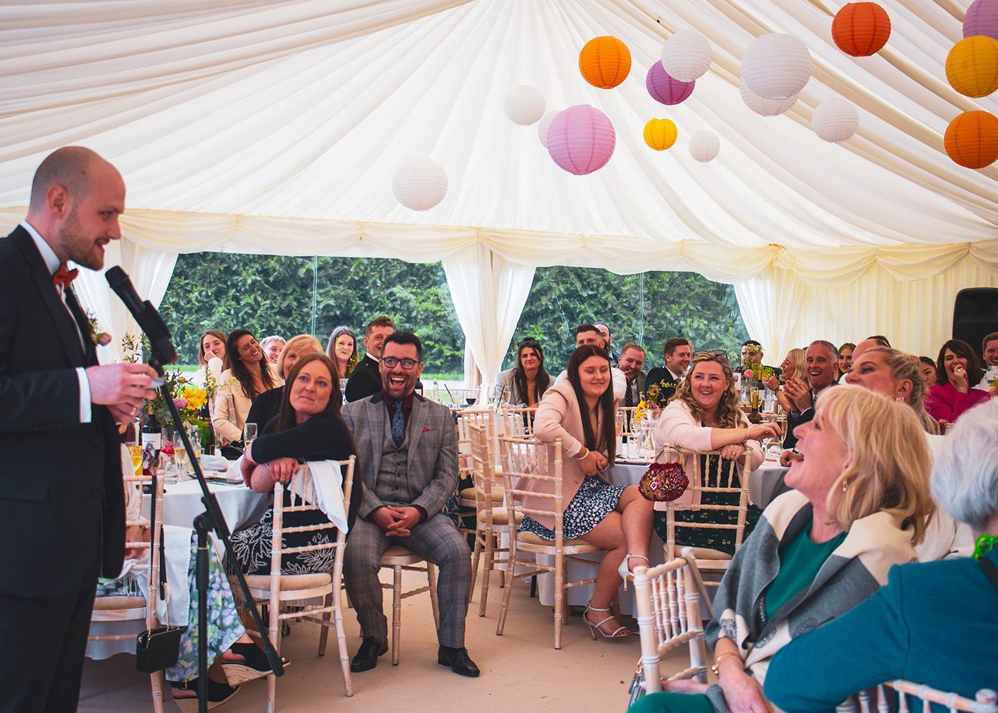A man is giving a speech at a wedding reception in a tent.