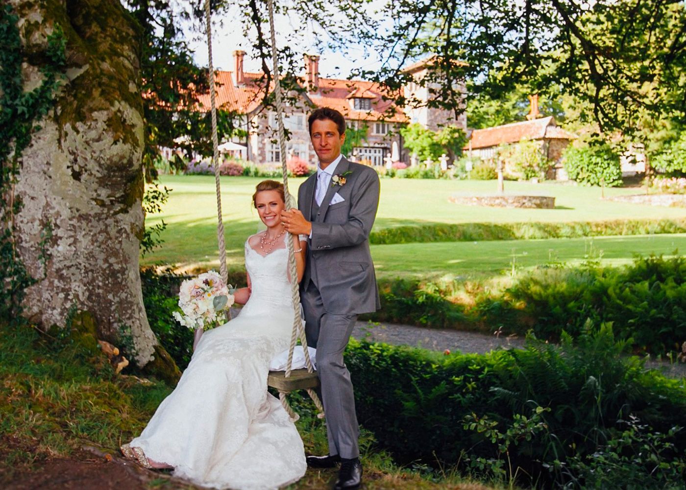 A bride and groom are posing for a picture while the bride is sitting on a swing.