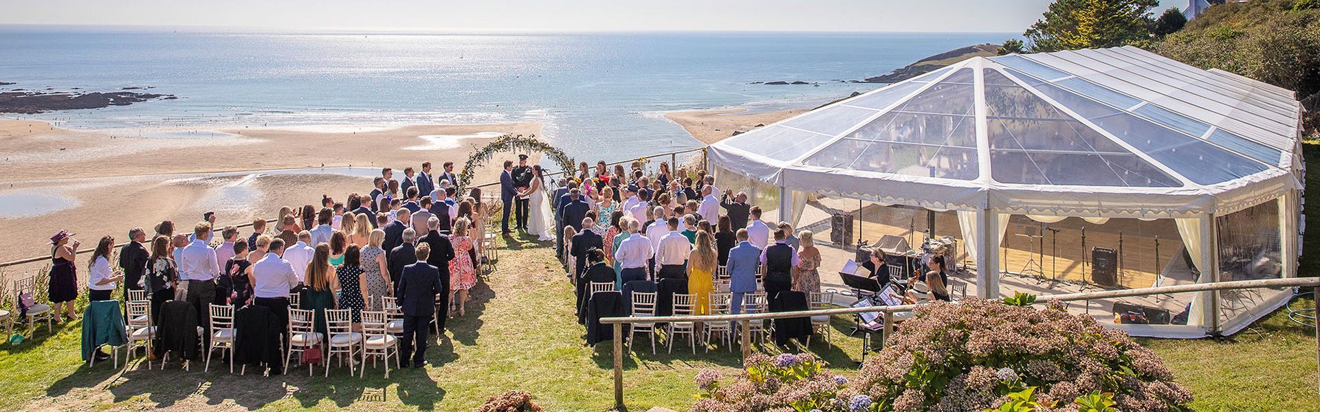 A large group of people are standing under a clear tent on the beach.