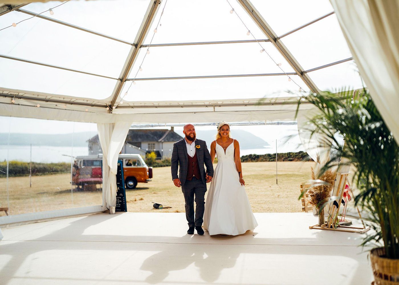 A bride and groom are walking through a clear tent holding hands.