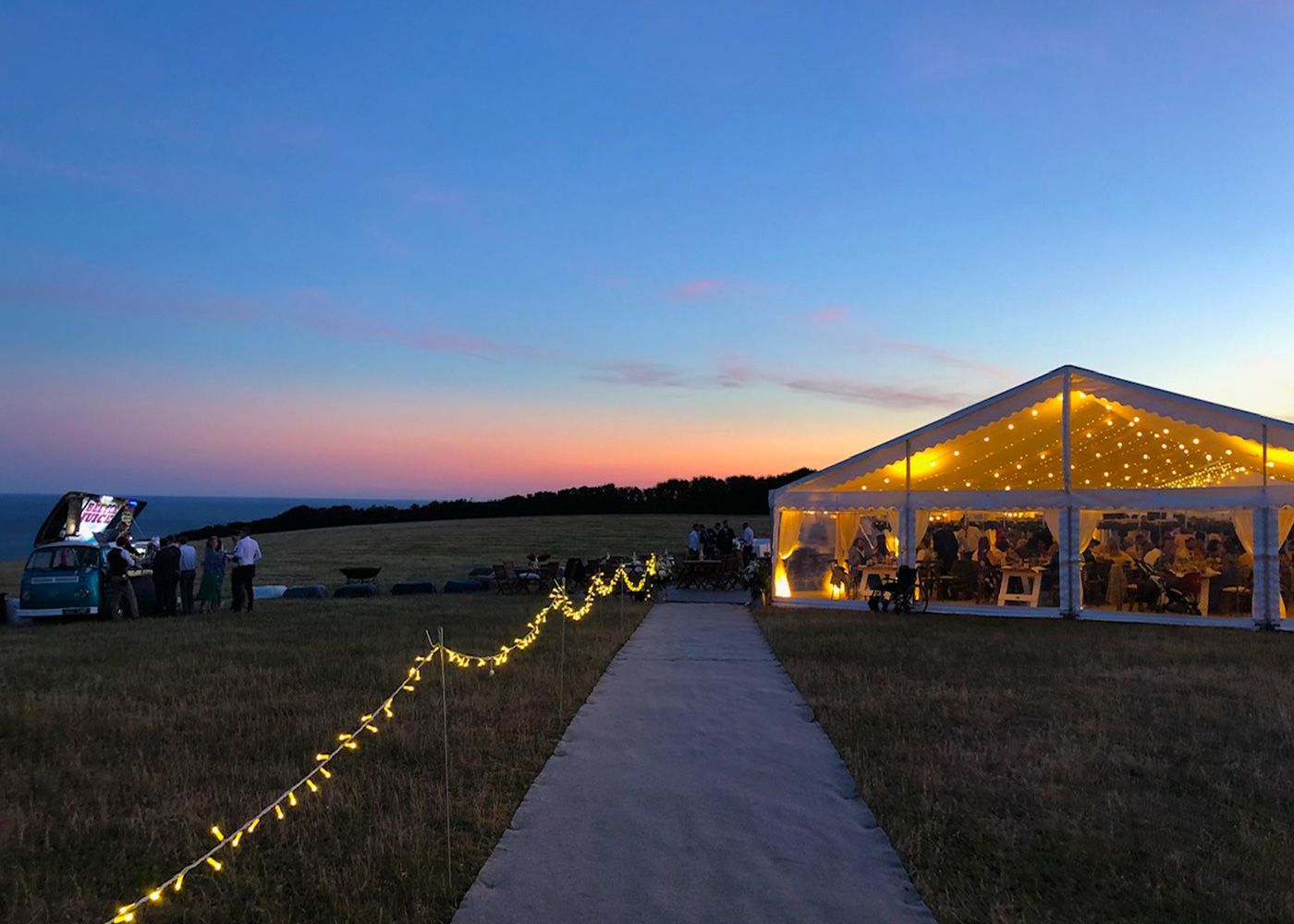A tent is lit up at night in a field with a sunset in the background.