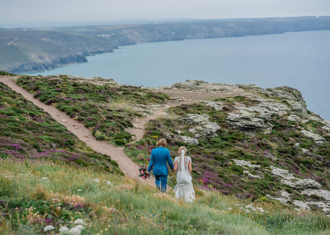 A bride and groom are walking down a path on a cliff overlooking the ocean.