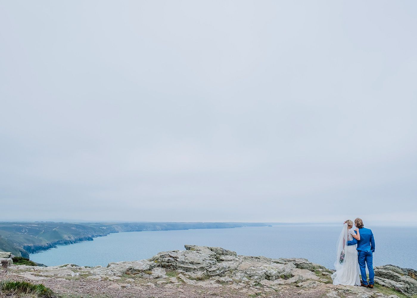 A bride and groom are standing on top of a hill overlooking the ocean.