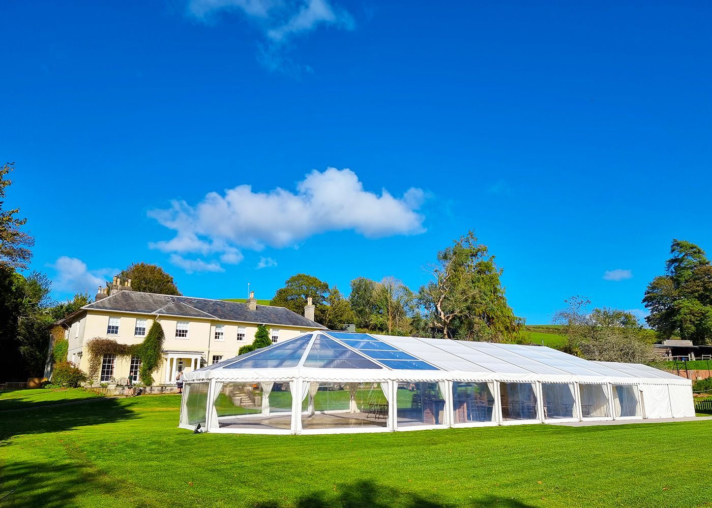 A large white tent is sitting in the grass in front of a large house.