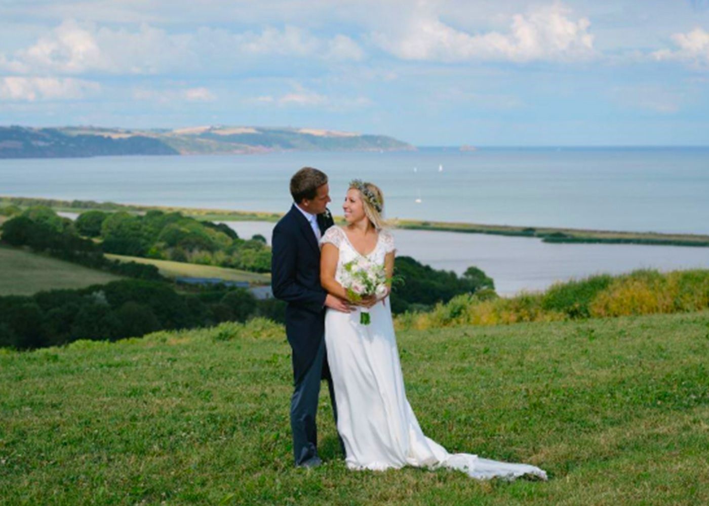 A bride and groom are standing on top of a grassy hill overlooking the ocean.