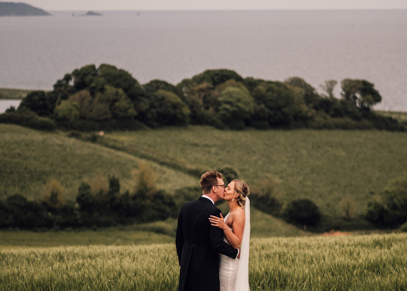 A bride and groom are kissing in a field with the ocean in the background.