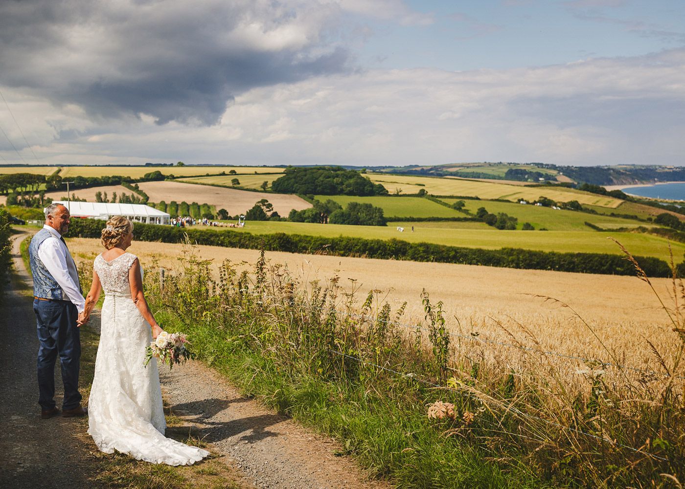 A bride and groom are standing on a dirt road looking at a field.