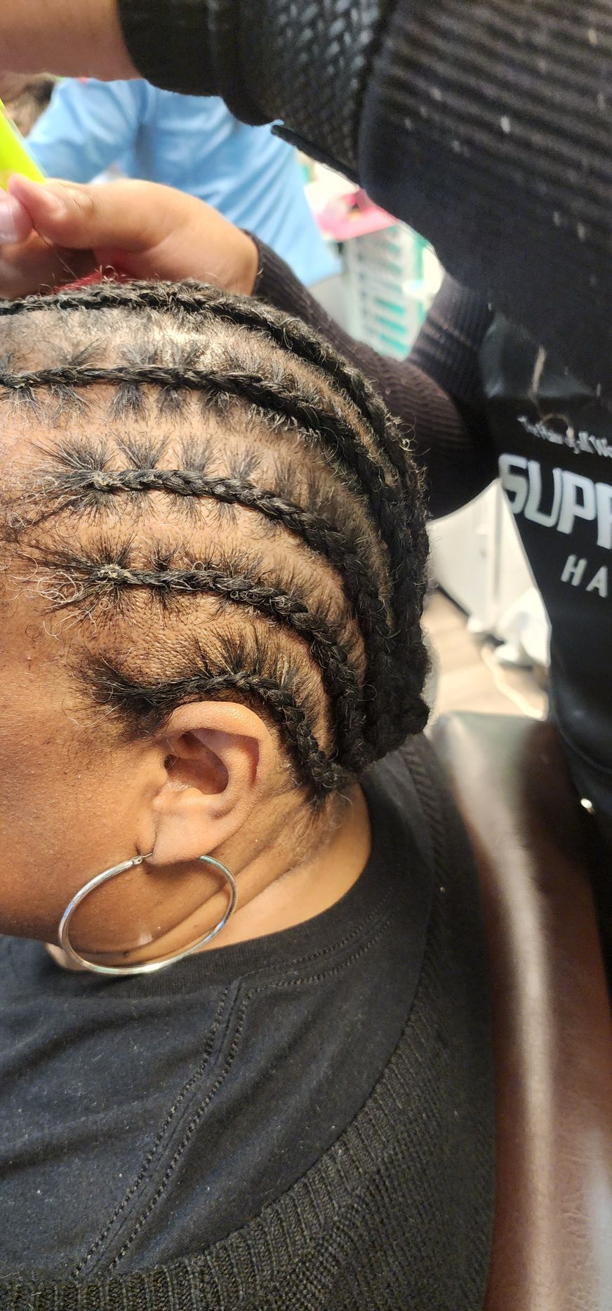 Person getting hair cornrowed by another person. Close-up of braided hair, ear and hoop earring visible.