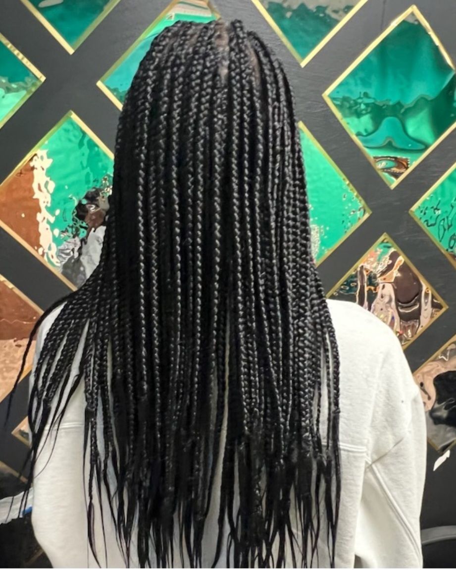 Woman with long black box braids, back view. Wearing white sweatshirt, standing in front of patterned wall.