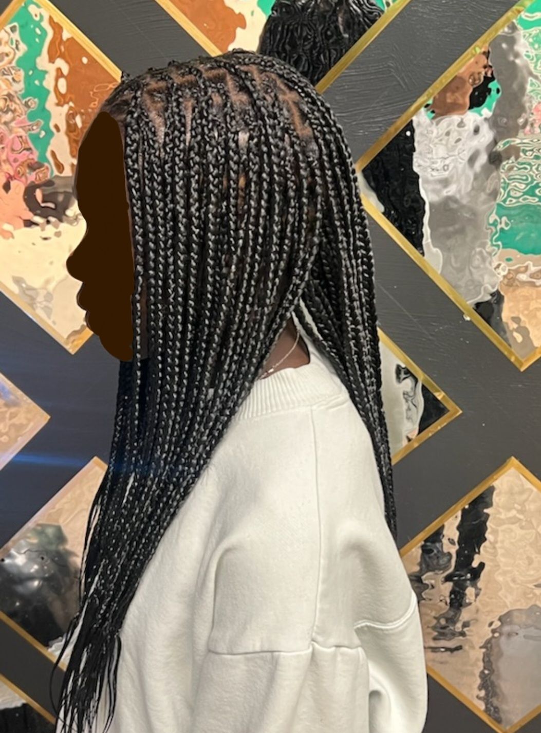 Side view of a person with long, thin black braided hair, wearing a white sweater, in front of an abstract art piece.
