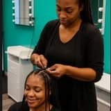 Woman braids another woman's hair in a salon with turquoise walls, both smiling.