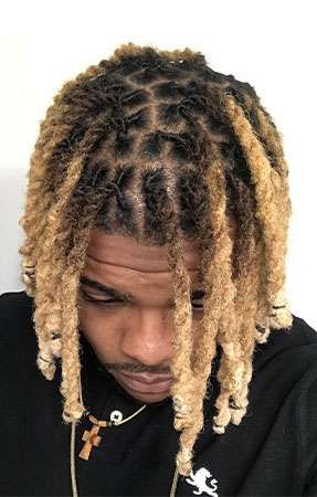 Man with blonde and black locs, wearing a necklace, looking down.