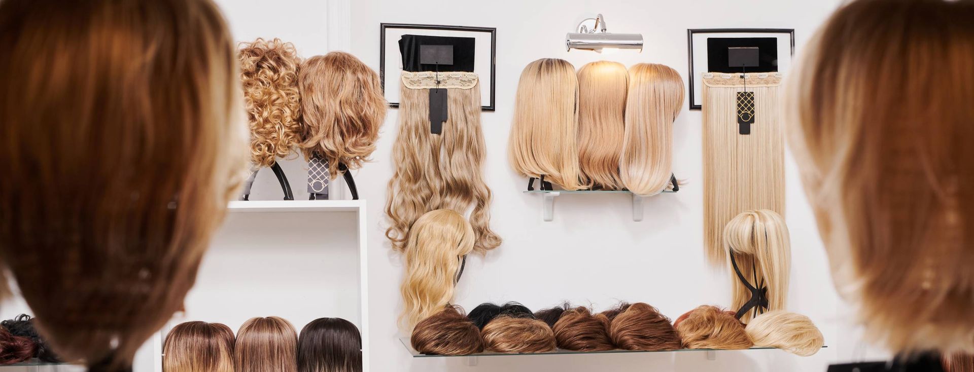 Wigs on display in a shop, including brown, blonde, and curly styles, against a white wall.