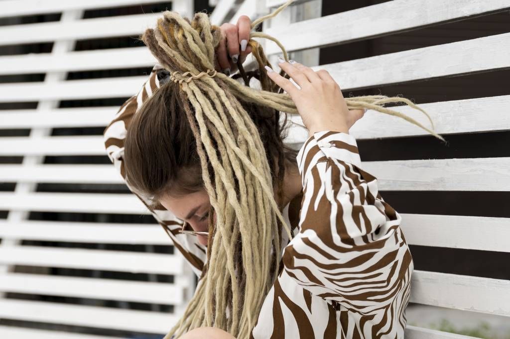 Woman with blonde dreadlocks, wearing a zebra print shirt, tying her hair up against a white slatted backdrop.