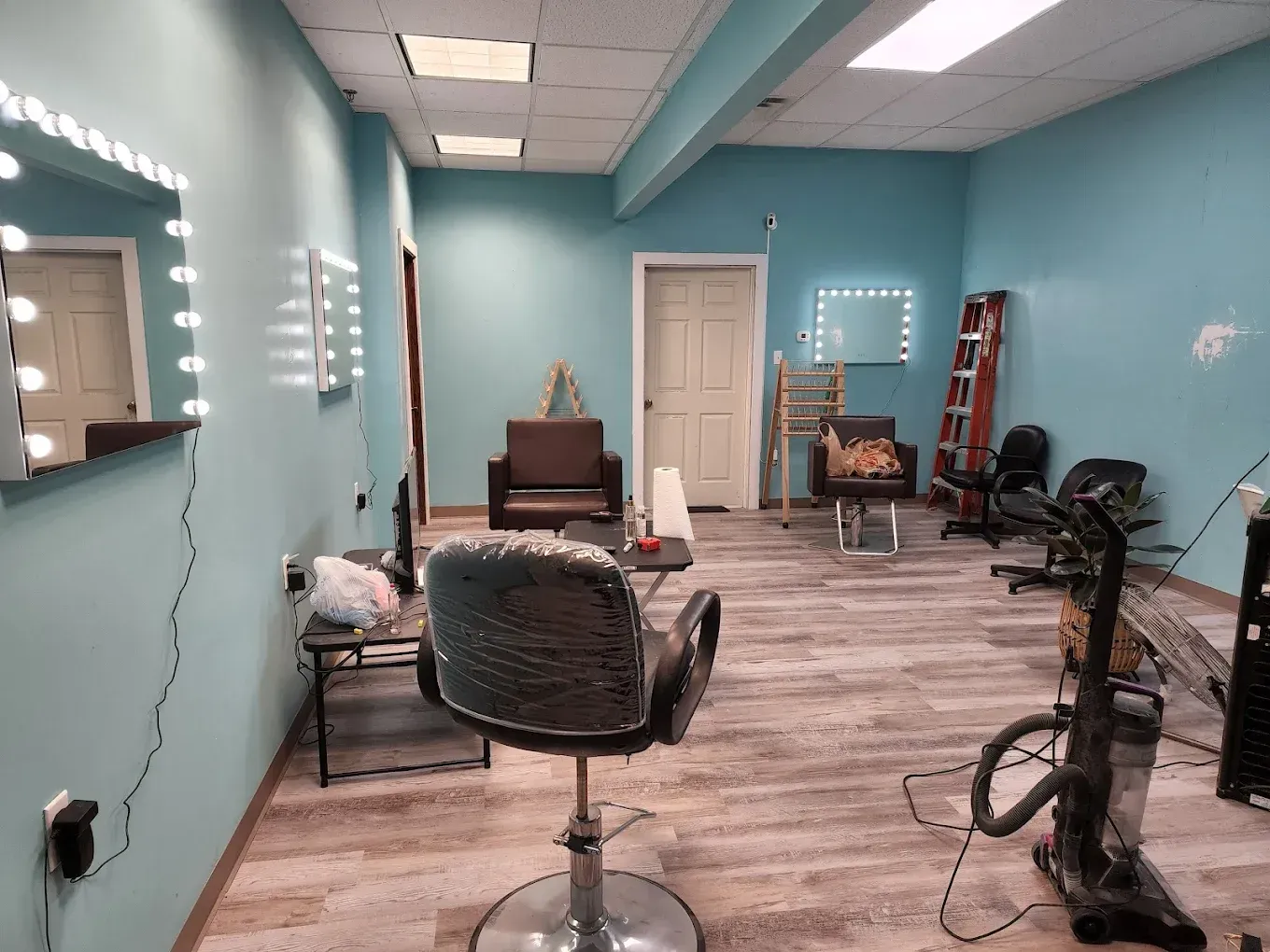 Hair salon interior, turquoise walls, three illuminated mirrors, stylist chair, vacuum cleaner, brown wooden floor.