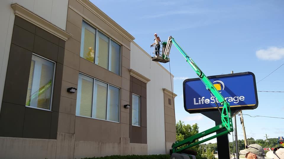 Two workers on a green lift painting exterior of a Life Storage building with a blue sign.