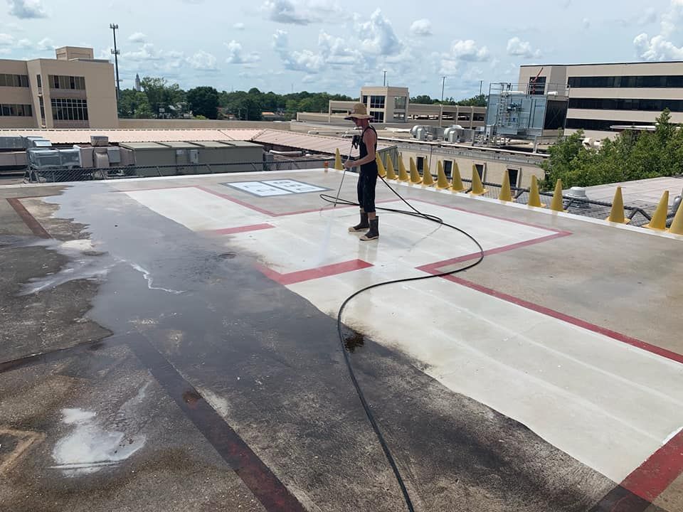 Person pressure washing a rooftop helipad, with buildings and sky visible.