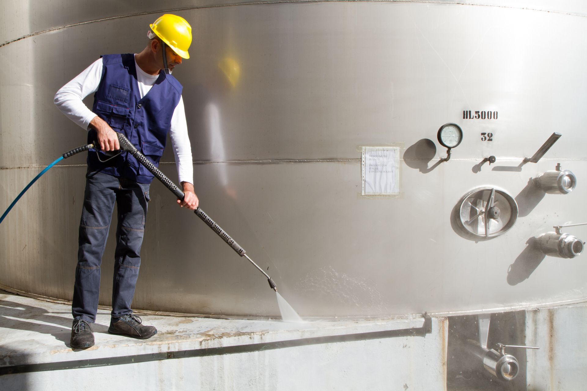 Man in safety gear power washing a large metal industrial tank.