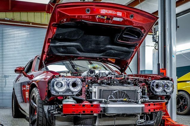 A red car with the hood up is being worked on in a garage.