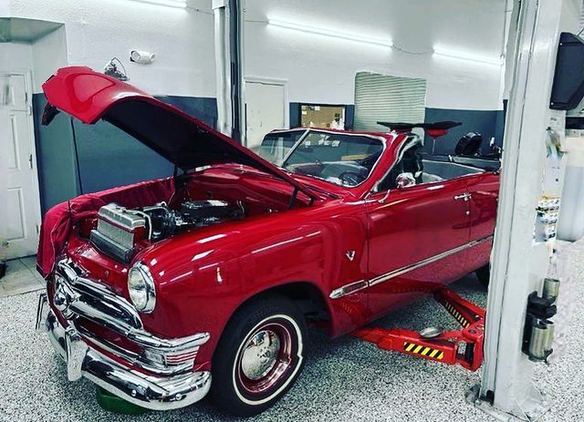 A red car with the hood up is on a lift in a garage.
