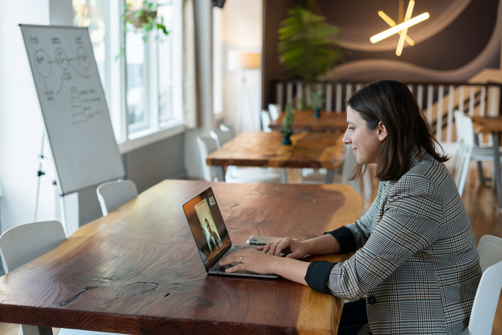 Woman in blazer on a video call at a wooden table in a well-lit room. A whiteboard is to the left.