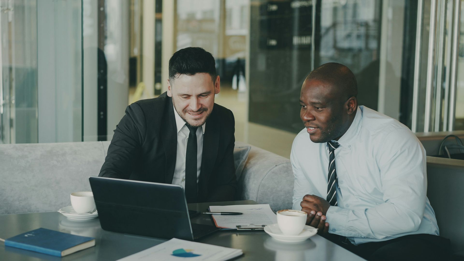 Two men in suits, smiling, looking at a laptop, sitting at a table with coffee cups and documents.
