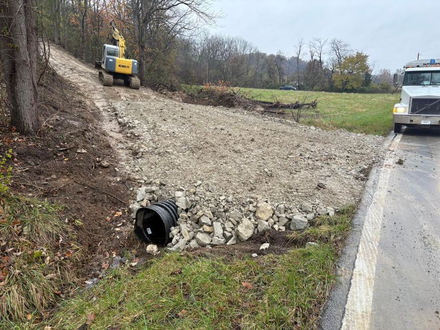 Construction site: excavator on dirt path above gravel-covered slope, culvert pipe, and truck parked on road.