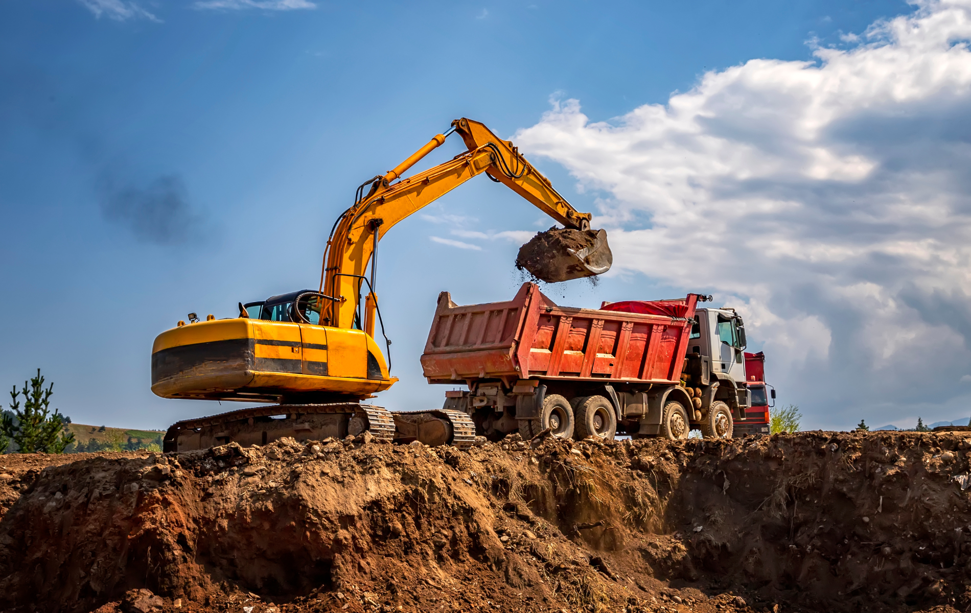 Yellow excavator loading a red dump truck with dirt on a construction site, under a cloudy blue sky.