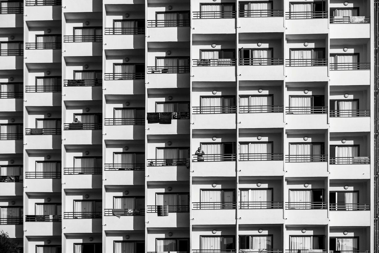 Black and white photo of a multistory apartment building with rows of balconies, casting shadows.