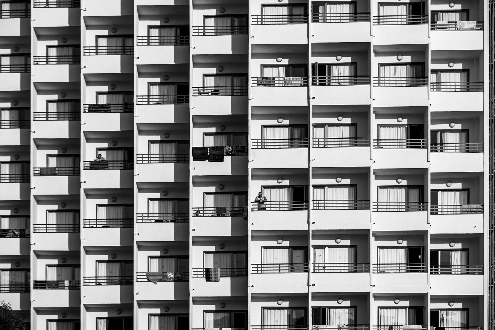 Black and white photo of a multistory apartment building with rows of balconies, casting shadows.