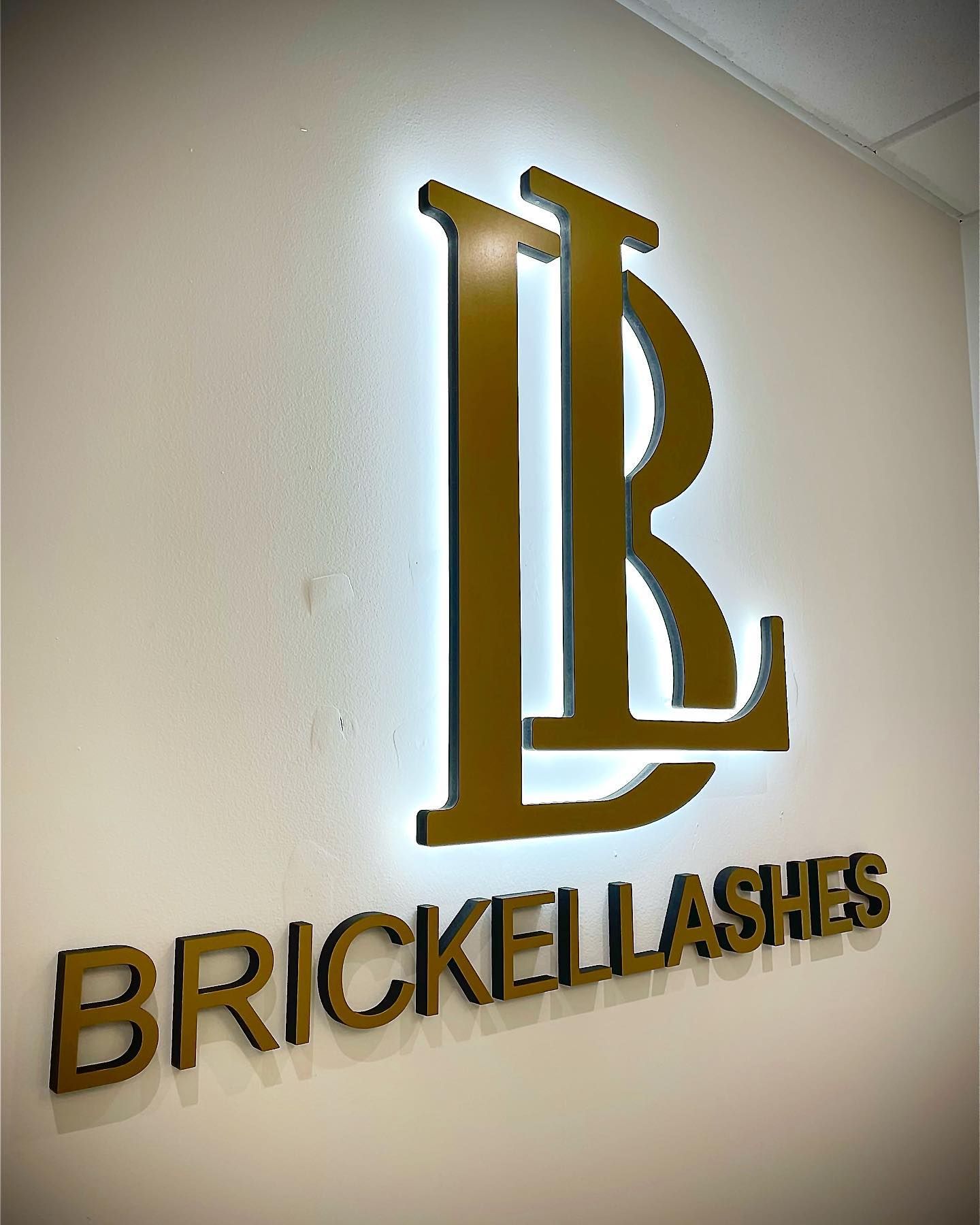 A gold logo for brickellashes is on a white wall.