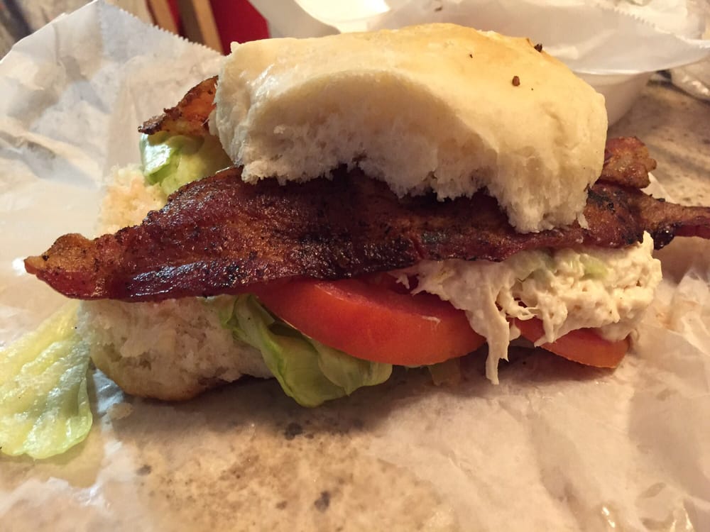 BLT sandwich on a roll with visible bacon, tomato, lettuce, and chicken salad on white paper.
