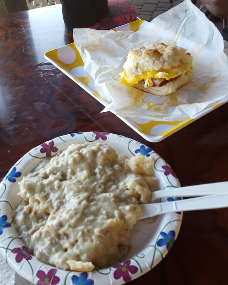 Biscuit sandwich on a yellow plate, gravy with utensils on a floral paper plate, on a wooden table.
