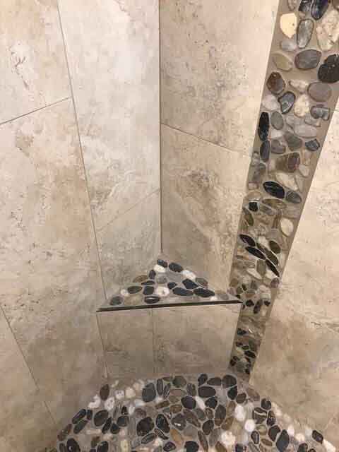 A shower with a corner shelf and a tiled floor with rocks.